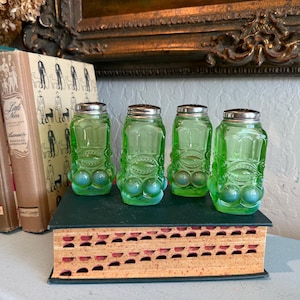 May include: Four vintage green glass salt and pepper shakers with silver lids. The shakers have a unique design with a face-like shape and three blue glass balls at the base. They are displayed on a decorative book.