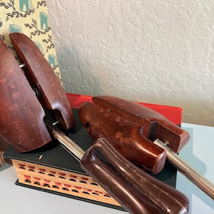 May include: Two brown wooden shoe trees with metal rods. The shoe trees are resting on a book with a red and white patterned cover.