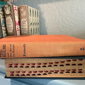 May include: An orange hardcover book titled "The House and The Art of its Design" by Kennedy. The book is stacked on top of a smaller, brown book with a patterned cover.