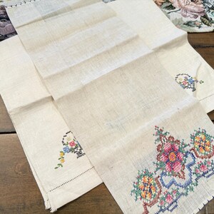 May include: Two white linen tea towels with embroidered floral designs. One towel has a single floral design in the centre, while the other has a larger floral design in the bottom right corner.