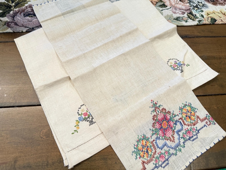 May include: Two white linen tea towels with embroidered floral designs. One towel has a single floral design in the centre, while the other has a larger floral design in the bottom right corner.