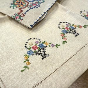 May include: Set of three vintage linen tea towels featuring embroidered floral baskets in shades of blue, pink, yellow, and green. The towels have a scalloped edge and are made from soft, natural linen.