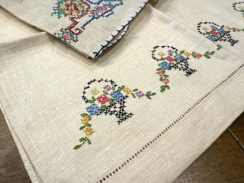 May include: Set of three vintage linen tea towels featuring embroidered floral baskets in shades of blue, pink, yellow, and green. The towels have a scalloped edge and are made from soft, natural linen.