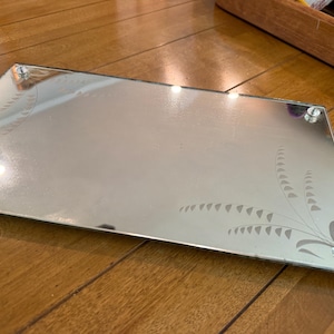 May include: A rectangular mirrored vanity tray featuring a reflective silver surface with etched floral patterns in the corners. The tray is supported by four small, clear, spherical glass feet positioned at each corner of the base.