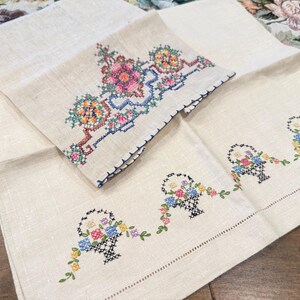 May include: Two embroidered linen tea towels. One towel features a floral design with a basket of flowers. The other towel has three baskets of flowers with trailing vines.