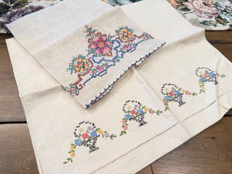 May include: Two embroidered linen tea towels. One towel features a floral design with a basket of flowers. The other towel has three baskets of flowers with trailing vines.