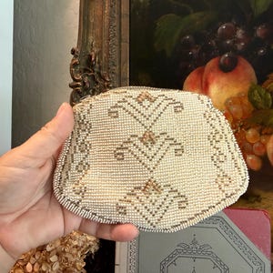 May include: A small, rectangular coin purse covered in tiny white beads with a geometric pattern created by metallic silver and copper-colored beads. The purse features a structured frame and a repeating symmetrical design across the front surface.