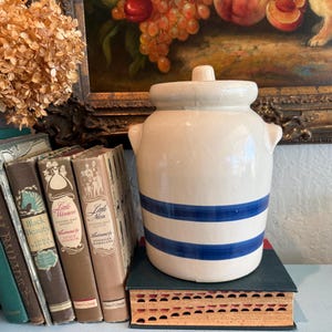May include: A cream-colored stoneware crock with a lid and two blue stripes. The vintage jar has small handles and sits on a stack of books. A collection of antique books and a painting are in the background.