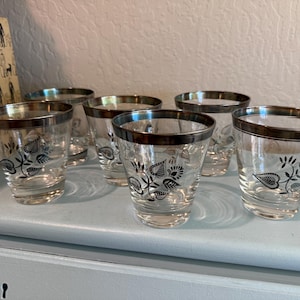 May include: Set of six vintage clear glass tumblers with silver rims and a black floral design. The glasses are arranged on a light blue surface, showcasing their elegant design. These glasses are perfect for serving beverages.