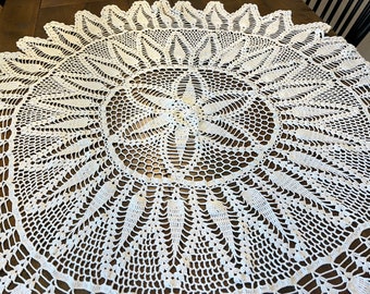 May include: A white crocheted tablecloth with a delicate floral pattern. The tablecloth is round and has a scalloped edge.