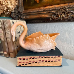 May include: A peach-colored swan-shaped decorative dish. The swan is perched on a dark green book with a decorative edge. A collection of antique books is visible in the background, along with a framed painting.