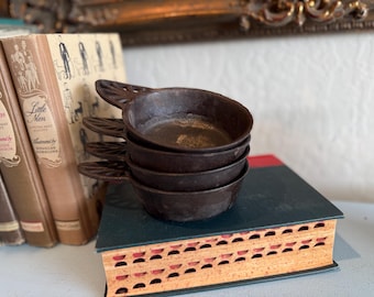 May include: A stack of four small, dark brown cast iron pans with decorative handles. The pans are stacked on top of a vintage book with a red and black cover, next to a stack of antique books.