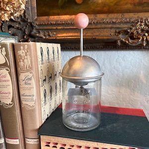 May include: A vintage glass and metal kitchen tool with a pink wooden knob. The clear glass jar has measurements printed on the side. The tool is sitting on a stack of books, including "Little Women" and "Little Men".