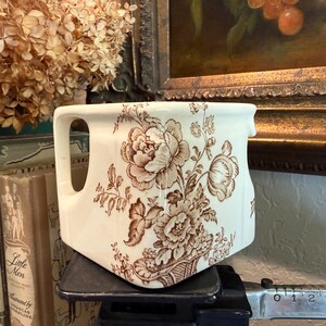 May include: A cream-colored ceramic creamer featuring a brown floral transferware pattern of peonies and foliage. The vessel has a square-shaped body with a single handle and a small pouring spout, positioned on a scale with a visible metric ruler.