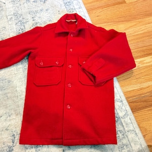 May include: A bright red wool jacket with a collared neckline, two chest pockets, and button closures. The jacket has long sleeves with buttoned cuffs. The garment is laid out on a patterned rug and wooden floor.