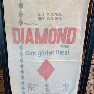 May include: A vintage Diamond Brand corn gluten meal sack. The off-white sack features red text that reads "DIAMOND BRAND" and "corn gluten meal." It also includes the text "100 POUNDS NET WEIGHT" and a red diamond graphic. The sack has a series of gray diamond shapes down the side.