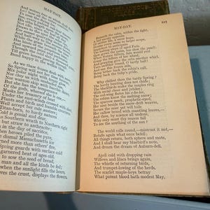 May include: Open book with aged, yellowed pages. The left page displays the title "MAY-DAY" and a poem. The right page also features the title "MAY-DAY" and a continuation of the poem. The book is open on a light blue surface.