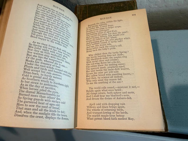 May include: Open book with aged, yellowed pages. The left page displays the title "MAY-DAY" and a poem. The right page also features the title "MAY-DAY" and a continuation of the poem. The book is open on a light blue surface.