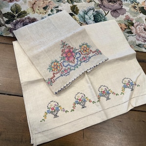 May include: Two white linen tea towels with embroidered floral designs. One towel features a single floral design with a basket of flowers, while the other towel has a repeating pattern of baskets of flowers.