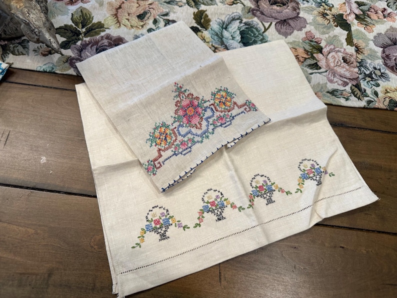 May include: Two white linen tea towels with embroidered floral designs. One towel features a single floral design with a basket of flowers, while the other towel has a repeating pattern of baskets of flowers.
