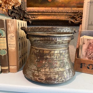 May include: A weathered, metallic bucket with a wide, flared base and a tapered neck. The surface features a distressed, tarnished finish with visible signs of wear. A sturdy, textured metal handle with a cross-hatched pattern is attached to the top rim.