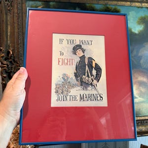 May include: A framed vintage recruitment poster featuring a person in a dark military uniform with the text IF YOU WANT TO FIGHT! JOIN THE MARINES. The print is set against a bright red matting inside a thin blue frame, displaying a historical illustration.