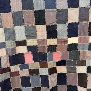 May include: Close-up of a patchwork quilt with a checkerboard pattern. The quilt is made of various fabrics in shades of blue, gray, brown, and pink. The squares are stitched together with visible seams, creating a textured surface.