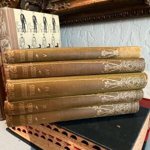 May include: A stack of antique books with aged, tan covers. The spines display the title "Emerson's Works" in gold lettering, with decorative floral patterns. The books are numbered I through V. Other books are visible in the background.