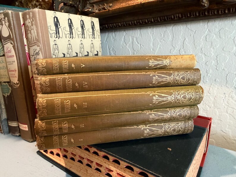 May include: A stack of antique books with aged, tan covers. The spines display the title "Emerson's Works" in gold lettering, with decorative floral patterns. The books are numbered I through V. Other books are visible in the background.