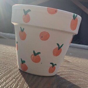 May include: White terracotta flower pot with a hand-painted design of orange fruit with green leaves.