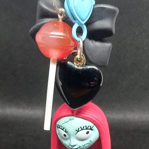 May include: A keychain featuring a Sally figure from The Nightmare Before Christmas, a black heart charm, a red lollipop, and a blue heart-shaped clasp. The keychain has a purple clasp and a black bow.