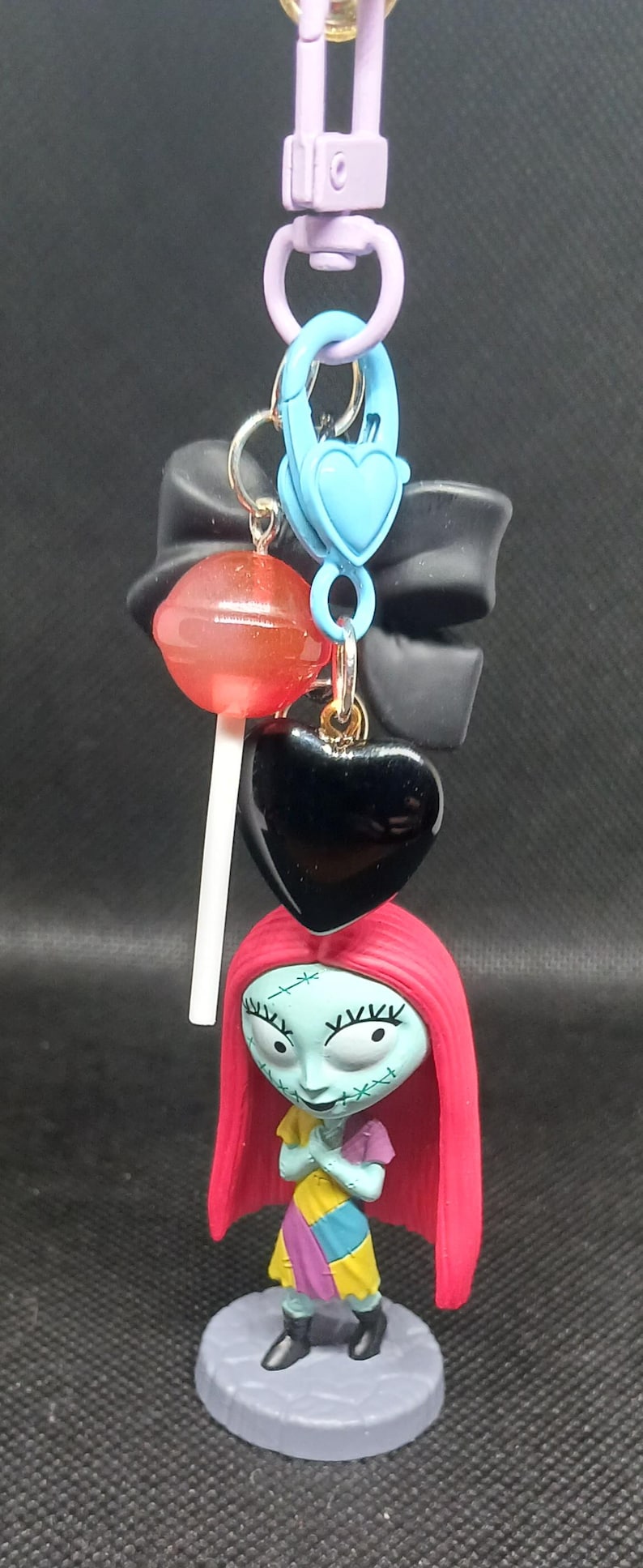 May include: A keychain featuring a Sally figure from The Nightmare Before Christmas, a black heart charm, a red lollipop, and a blue heart-shaped clasp. The keychain has a purple clasp and a black bow.