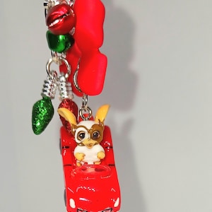 May include: A red car charm with a Gremlin figure wearing goggles. The charm is attached to a keychain with red and green bells, red shapes, and green glittery light bulb charms. The keychain is silver.