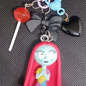 May include: A collection of keychains and charms, including a Sally figure with red hair and a patchwork dress, a black bow, a heart, a lollipop, and various metal clasps and rings. The items are in various colors.