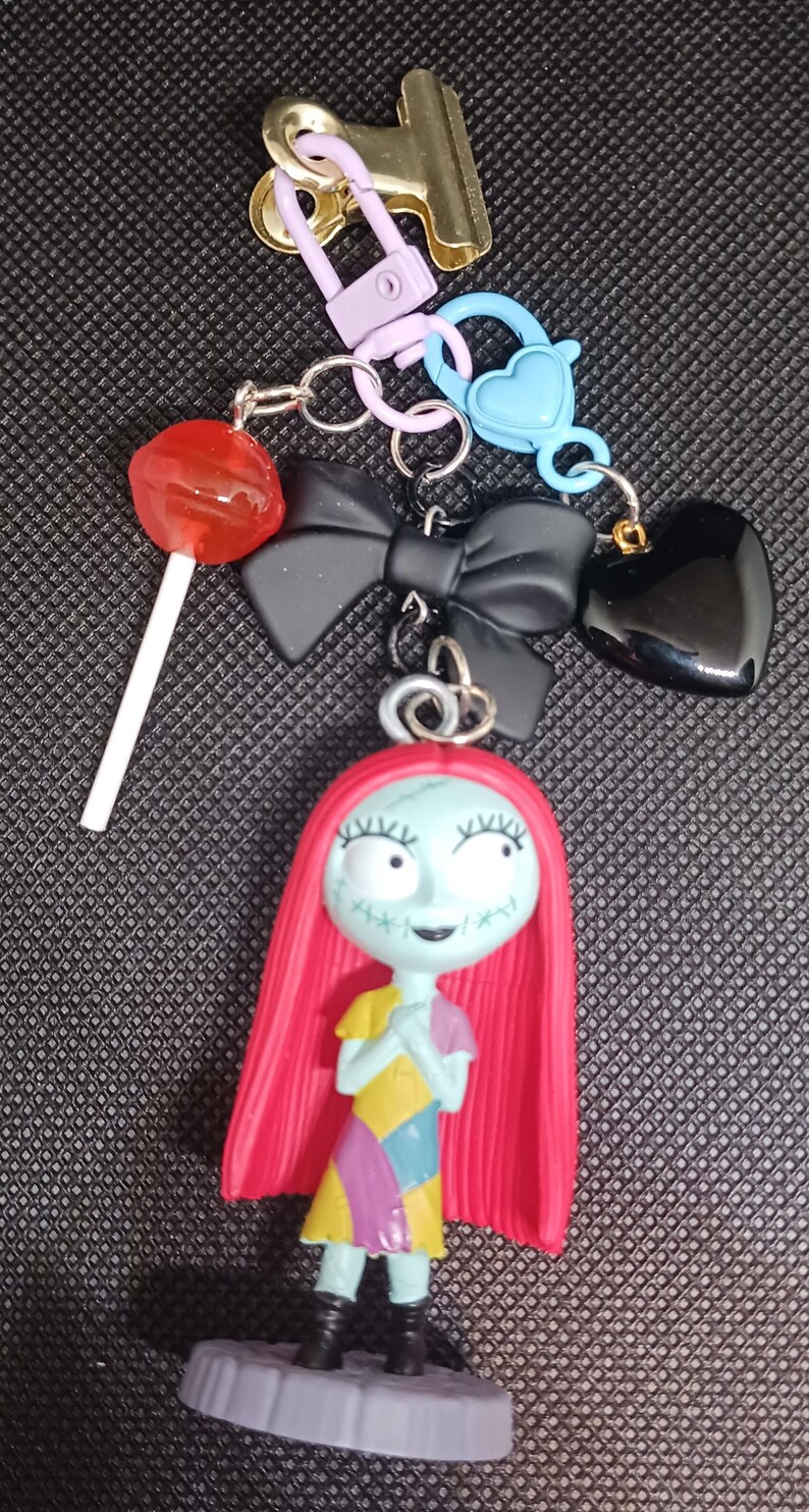 May include: A collection of keychains and charms, including a Sally figure with red hair and a patchwork dress, a black bow, a heart, a lollipop, and various metal clasps and rings. The items are in various colors.