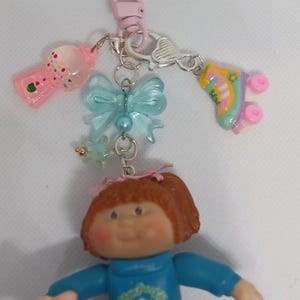 May include: A keychain featuring a Cabbage Patch Kids doll and various charms. The charms include a pink gumball machine, a light blue bow, and a roller skate. The doll is wearing a blue outfit and holding a small, white object.