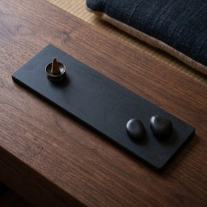 May include: A black rectangular incense holder with a small bronze incense cone and two smooth black stones. The holder sits on a dark wooden surface, suggesting a minimalist aesthetic.
