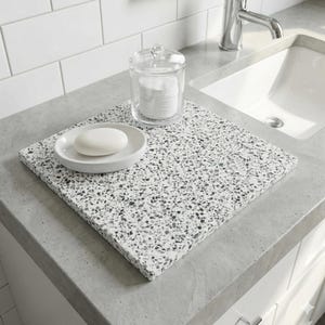 May include: A bathroom countertop with a square, speckled, gray and white decorative tray. A white soap dish with a bar of soap and a clear glass jar with cotton pads sit on the tray. A white sink and chrome faucet are in the background.