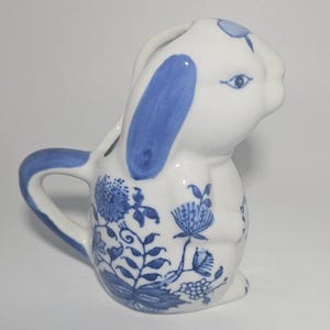 May include: A white ceramic bunny-shaped pitcher with blue accents. The pitcher features blue floral patterns and a handle. The bunny's ears and a small detail on its head are painted blue. The pitcher is designed to resemble a rabbit.