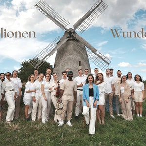 Puede incluir: Un grupo de personas con ropa blanca se encuentran frente a un molino de viento con un cielo azul de fondo. El texto "100 Millionen Wunder" está escrito en letras marrones en la parte superior de la imagen.