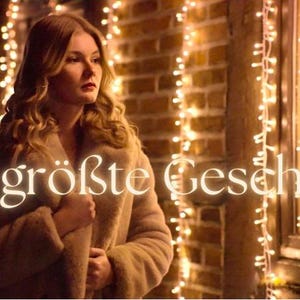 Könnte beinhalten: Eine Frau in einem beigen Mantel steht in der Nähe eines Fensters, das mit Lichterketten geschmückt ist. Der Text "Das größte Geschenk" ist in leuchtender Schrift dargestellt. Die Szene spielt nachts, mit warmer, einladender Atmosphäre.