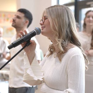 Könnte beinhalten: Eine Frau singt in ein Mikrofon auf einem Ständer, mit einer Band im Hintergrund. Sie trägt ein weißes, strukturiertes Langarmshirt und eine Brille. Weitere Bandmitglieder sind ein Gitarrist, ein Schlagzeuger und zwei Frauen.
