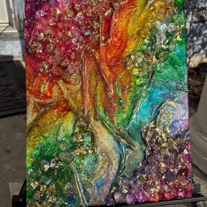 May include: Abstract resin art piece with a rainbow color scheme, featuring red, orange, yellow, green, blue, and purple hues. The artwork incorporates gold leaf accents and a textured surface, mounted on a black easel.