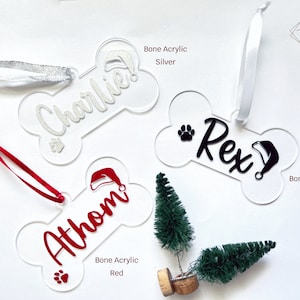 May include: Three bone-shaped Christmas ornaments with the names "Charlie", "Rex", and "Athon" on them. The ornaments are made of clear acrylic and have a silver, black, and red finish. Each ornament has a paw print and a Santa hat.