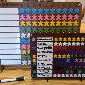 May include: A wooden board with a white dry-erase surface and a grid of coloured meeples. The board is labelled "Mallory & Steve" and has numbers 1 through 10. The meeples are arranged in rows of different colours, including blue, pink, yellow, and green. A second wooden board with a similar grid of meeples is also visible. The board is labelled with the names of board games, including "Carcassonne", "Manabi", "Tranquility", "Cartographers", "Seafall", "Barenpark", "King's Dilemma", "Space Base", "Legacy of Yu", and "Star Realms".