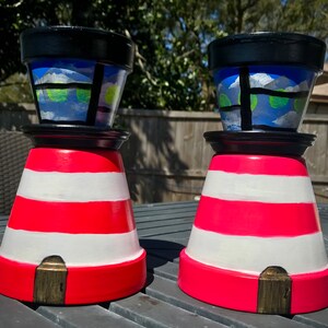 May include: Two painted terracotta flower pots shaped like lighthouses. The pots are red and white striped with black tops and blue windows. The windows have white clouds and green circles painted on them.