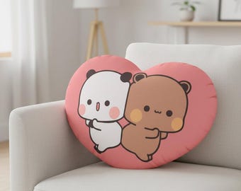 May include: Heart-shaped decorative pillow featuring a cartoon panda and bear embracing on a pink background. The panda is white with black ears, and the bear is brown with yellow cheeks. The pillow is on a white sofa.
