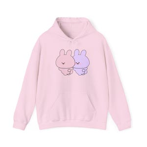 May include: A pink hoodie with a graphic of two cartoon bunnies holding hands. The bunny on the left is pink and the bunny on the right is purple.