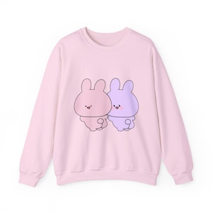 May include: A light pink sweatshirt featuring a cartoon illustration of two bunnies. One bunny is pink, and the other is purple. The bunnies are drawn with black outlines and have small, smiling faces. The sweatshirt has long sleeves and a crew neck.