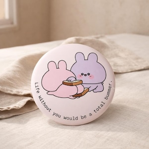 May include: A round, light pink compact mirror with a cartoon illustration of two bunnies sharing toast. The text "Life without you would be a total bummer" is printed around the edge of the mirror.